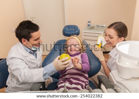 woman holding mirror and man giving apple to girl. dental office and little girl patient sitting in chair