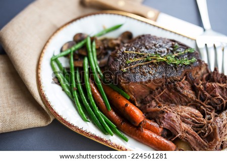 Tender roast beef with carrots and green beans on a white porcelain platter with a gold rim and serving fork against a gray tablecloth.