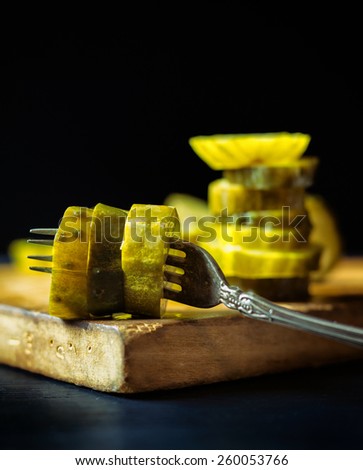 Stack of pickle slices behind an antique fork with more pickles on a rustic wooden cutting board.