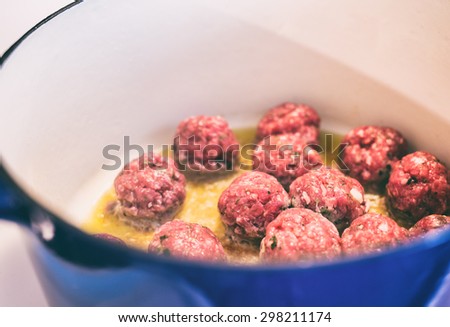 Fresh meatballs frying in olive oil on a stovetop in a blue dutch oven.  Vintage filter with grain applied.