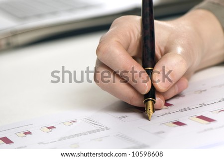 close up of a hand holding a pen above some papers with graphics. Laptop on the background.