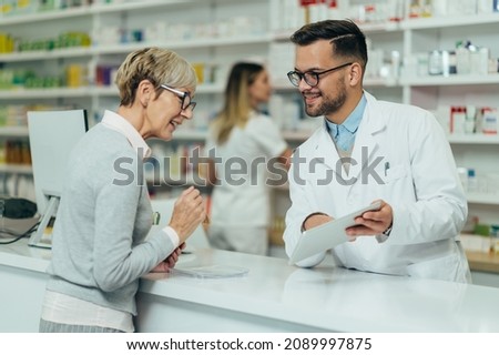 Young male pharmacist giving prescription medications to senior female customer in a pharmacy with female pharmacist in the background Imagine de stoc © 
