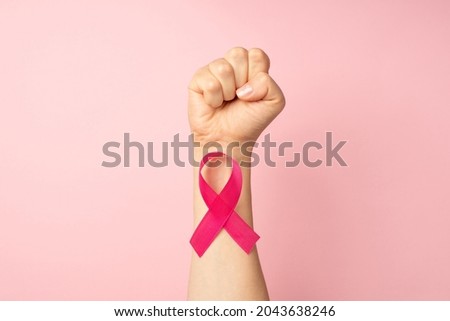First person top view photo of raised female hand with clenched fist and pink ribbon on wrist symbol of breast cancer awareness on isolated pastel pink background with copyspace