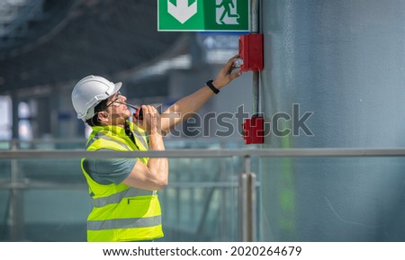 Engineer wearing safety unifrom and helmet under checking fire alarm emergency system in industry factory and exit door is factory security protection concept.