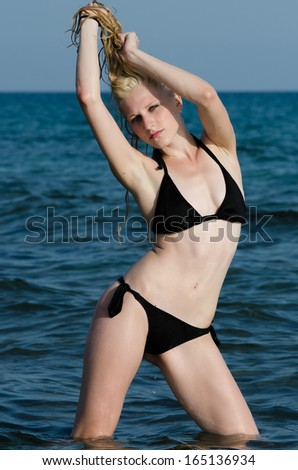 Sexy young blond woman posing in the sea in her black bikini, long blonde hair contrasting dark blue color of the sea