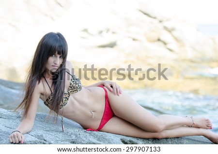 An attractive young woman lying on rocks at the beach. She is wearing a red and animal print striped bikini. Horizontal shot