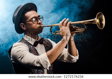male musician in hat and eyeglasses playing on trumpet on stage with dramatic lighting and smoke 商業照片 © 