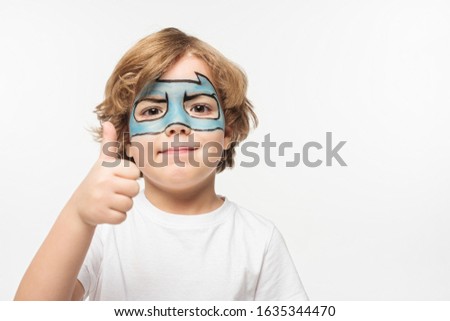 cheerful boy with mask painted on face showing thumb up while looking at camera isolated on white