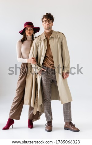 full length view of stylish woman in hat touching hand of trendy man in trench coat on grey