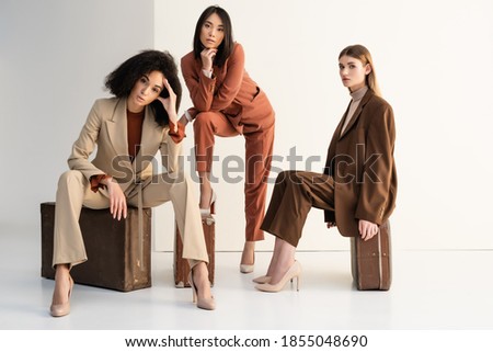 full length of stylish multicultural women in suits sitting on suitcases on white Foto d'archivio © 
