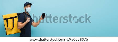 arabian delivery man in medical mask showing smartphone with blank screen on blue, banner