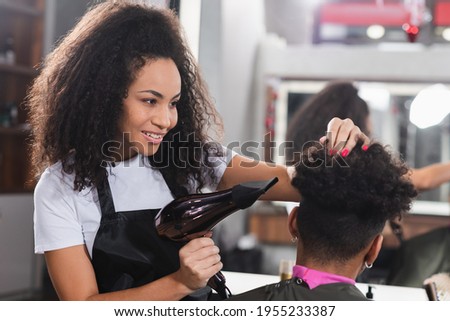 Positive african american hairdresser holding hair dryer near client on blurred foreground