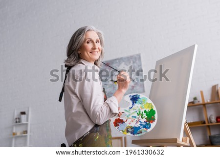 cheerful middle aged woman holding paintbrush and palette with colorful paints near blank canvas