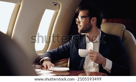 Smiling businessman holding cup and looking at window in private plane Imagine de stoc © 