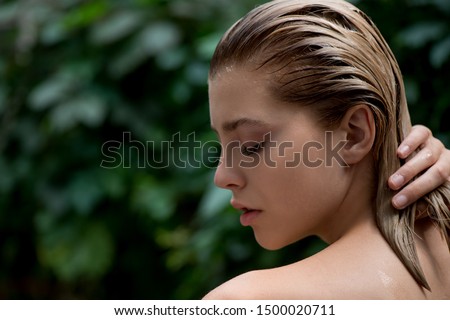 Beautiful young woman applies a balm, shampoo or conditioner to her damaged hair  front of plant tropical green leaves background.  SPA, bodycare, haircare  and skincare.  Close up, selective focus.