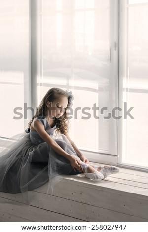 Little ballerina in gray dress puts on ballet shoes pointe front of the window.