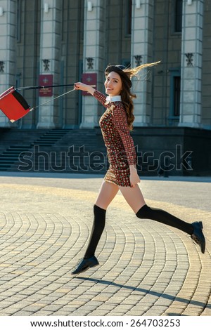 Beautiful brunette girl in stylish dress jumps on the city square. Fashion and city style. Facial expression. Various flags and the Orthodox Church on background.