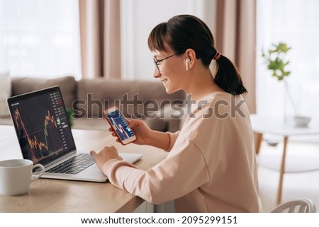 Smiling business woman trader analyst looking at laptop monitor, holding smartphone, wearing earphones. Investor broker analyzing indexes, trading online investment data on stock market graph at home Foto stock © 