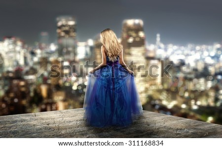 Beautiful blond girl in a blue cocktail dress is standing on the top of a skyscraper