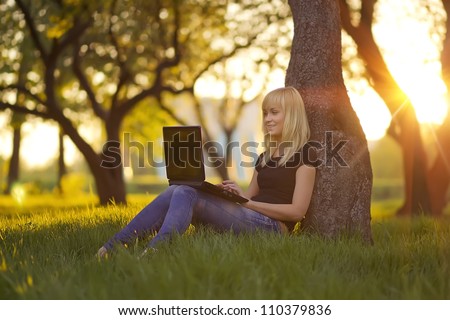Young woman with laptop sitting on green grass in the park at sunset