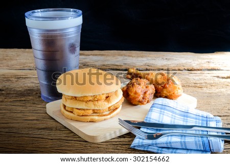 chicken hamburger and fried chicken, glass of cola on wooden cutting board with Knife and fork, napkin on top wooden table