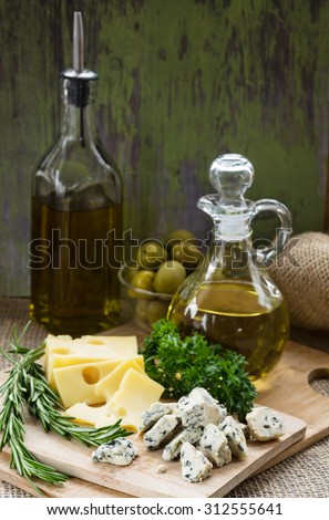 Cheese of two kinds on a cutting board, two bottles of olive oil, green olives in a glass bowl, and branches of rosemary and parsley.