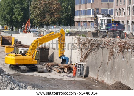 Excavator demolishing a reinforced concrete wall of a highway flyover while a worker wearing protective clothing and safety helmet is monitoring gas cylinders
