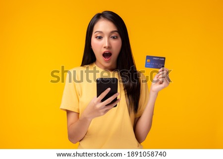 Happy young woman holding smart phone and showing credit card with surprise face Girl stand over yellow background, Online payment, hands holding credit card and using mobile phone for online shopping