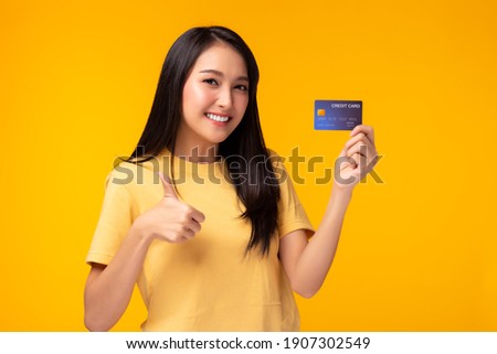 Young asian woman showing plastic credit card giving thumb up to credit card, standing over yellow background. Beautiful customer girl get satisfied of credit card service and promotion, smile face