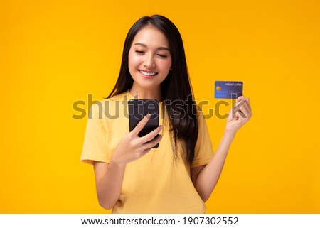Happy young woman using mobile phone showing plastic credit card standing over yellow background, Online payment, hands holding credit card and using smart phone for online shopping with smile face
