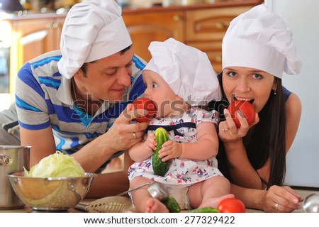 Young family with cooking hoods on their heads: a little cute baby-girl is sitting on the kitchen floor, she is eating a tomato - her parents are lying next to her, eating tomato and playing cooks