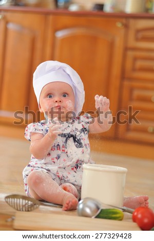 A little cute baby-girl with cooking hoods on her head is sitting on the kitchen floor, she is heavily soiled with wheat flour - she\'s playing cook