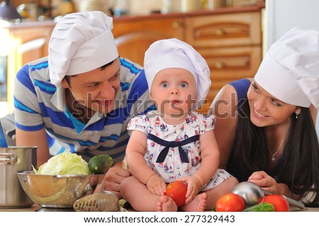 Young family with cooking hoods on their heads: a little cute baby-girl is sitting on the kitchen floor, she looks surprised - her parents are lying next to her smiling and playing cooks
