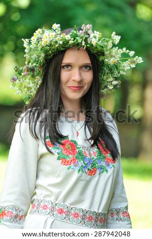Pretty smiling young woman\'s portrait outdoors: she is dressed in Ukrainian national decoration - embroidered flower-ornaments on her dress and with a wreath of fresh wildflowers on her head