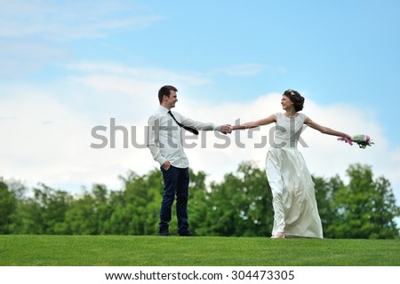 The bride pulls the hand of his beloved fiance and beckons to follow her - under their feet is luscious green grass and over their heads - bright blue sky. They are happy