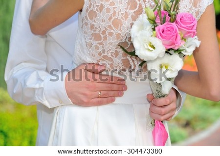 Male hands closeup: one hand holding a woman\'s waist in white festive dress, and another - a pink and white wedding bouquet.