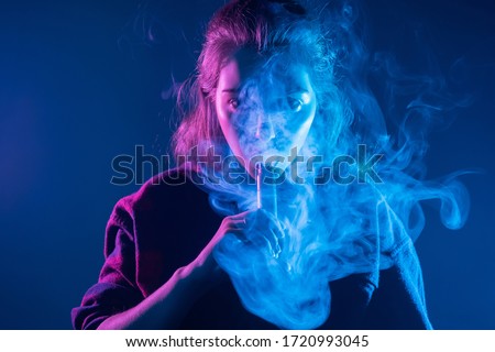 Smoking an electronic cigarette. The girl's face is covered with smoke from vape. Portrait of a female smoker in a cloud of smoke. A woman smokes a VAPE on a dark background.
