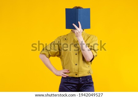 Woman covers her face with a book. Portrait of a girl without a face. Concept - selling books. Literature store. Girl with a book on a yellow background.  Woman reading a book while standing