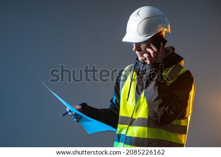 Builder with phone. Man in construction worker's uniform. Repairman in vest and helmet. Foreman on dark background. Builder calls customer. Portrait of builder with drawings. Foreman calls on phone Foto stock © 