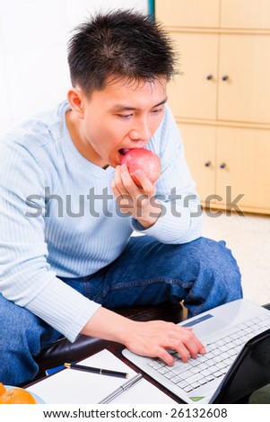 A young man eating apple while working on his laptop at home.