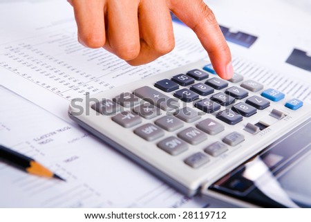 A finger is about to push the keypad in calculator, financial printouts are scattered around it.