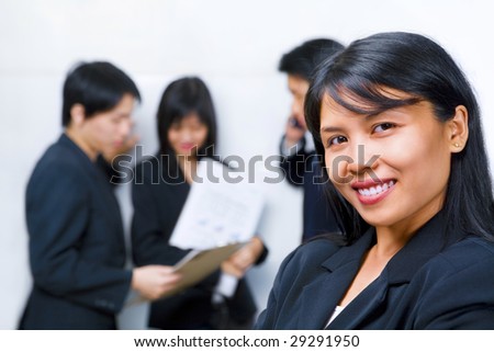 Young South East Asian Businesswoman smiling to camera with other people busy with each other on background