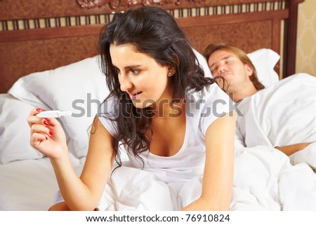 Lady looking at the pregnancy test device on bed while her spouse still sleeping