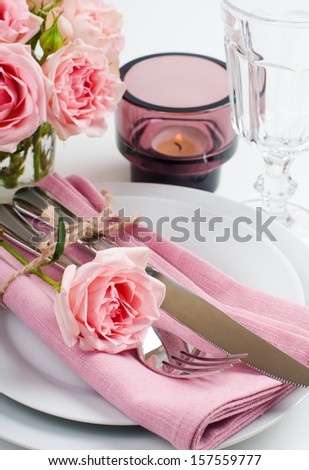 Beautiful festive table setting with roses, candles, shiny new cutlery and napkins on a white tablecloth.