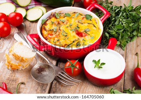 Vegetable casserole in a red pot with cheese, zucchini, cherry tomatoes, oregano and cream sauce on a wooden board, home cooking