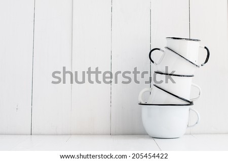 A stack of five white enameled mugs on painted wooden table, kitchen utensils and decor, rustic vintage kitchen background