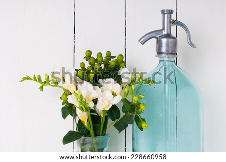 Vintage home decor, ancient turquoise siphon, freesias bouquet and bottles on a white wooden background.