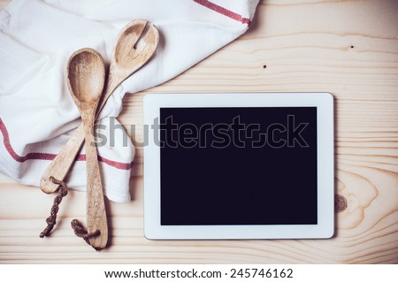 Tablet, towel and cooking utensils on wooden kitchen table, a blank screen.