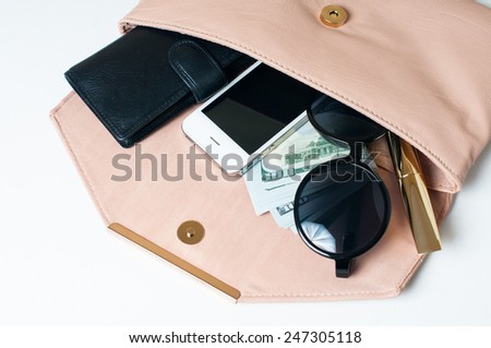 Cosmetics, sunglasses, money, purse and smartphone in an open beige woman\'s clutch handbag on a white background.