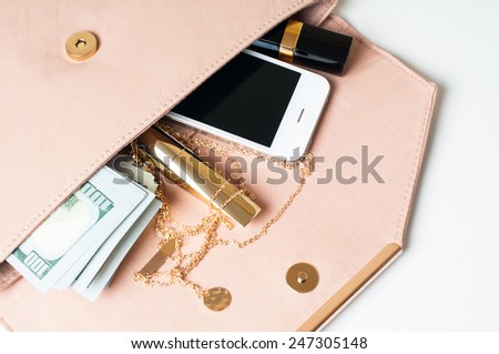 Cosmetics, jewelry, money and smartphone in an open beige woman\'s clutch handbag on a white background.